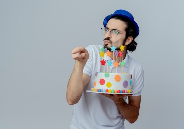 Guardando il lato confuso bell'uomo con gli occhiali e cappello blu tenendo la torta e punti a lato isolato su sfondo bianco con spazio di copia