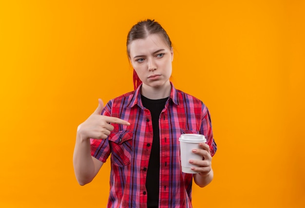 Guardando il lato giovane bella ragazza che indossa la camicia rossa punta il dito alla tazza di caffè in mano su sfondo giallo isolato