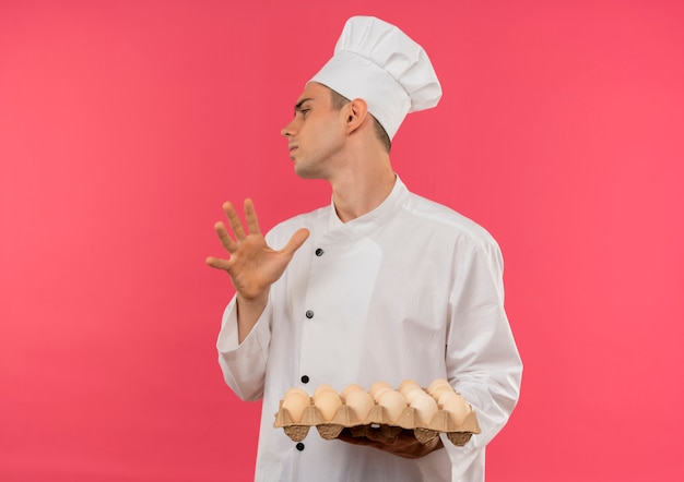 Guardando il lato giovane cuoco maschio che indossa l'uniforme del cuoco unico che tiene lotto di uova che mostrano il gesto di arresto sulla parete rosa isolata