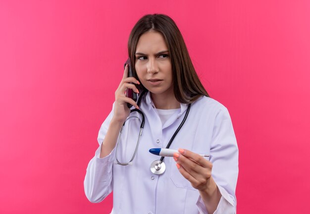 Guardando il lato giovane medico ragazza che indossa uno stetoscopio abito medico tenendo il termometro parla al telefono su sfondo rosa isolato