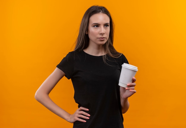 Guardando il lato giovane ragazza caucasica che indossa la maglietta nera tenendo la tazza di caffè mise la mano sul fianco isolato su sfondo arancione