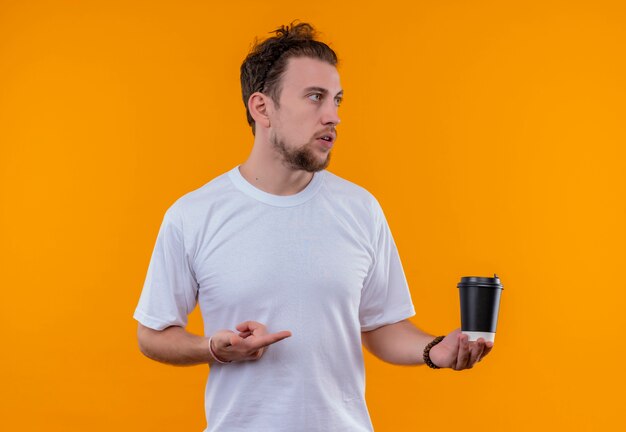 Guardando il lato giovane ragazzo che indossa la maglietta bianca indica la tazza di caffè sulla sua mano su sfondo arancione isolato