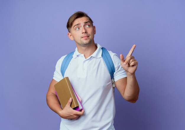 Guardando il lato giovane studente maschio bello che indossa la borsa posteriore in possesso di libri e punti a lato isolato sulla parete blu con lo spazio della copia