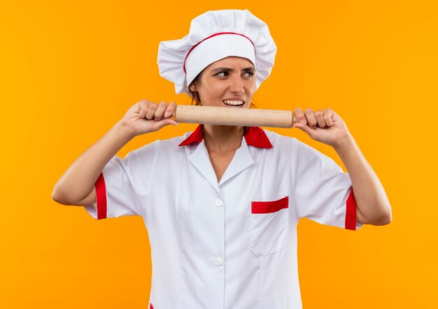 Guardando il lato interessato giovane cuoco femminile che indossa l'uniforme del cuoco unico che tiene il mattarello intorno alla bocca sulla parete gialla isolata con lo spazio della copia