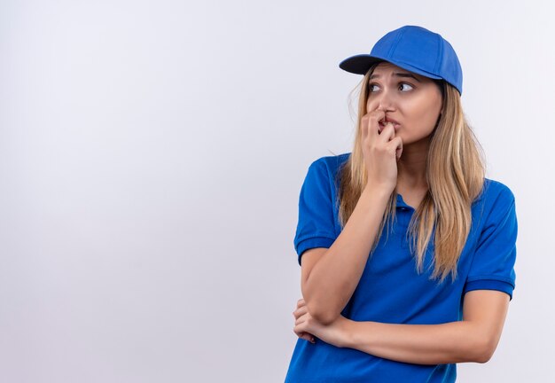 Guardando il lato interessato la giovane ragazza di consegna che indossa l'uniforme blu e il cappuccio che mette la mano sulla bocca isolata sulla parete bianca con lo spazio della copia