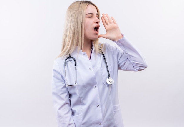 Guardando il lato medico giovane ragazza bionda che indossa stetoscopio e abito medico chiamando qualcuno sul muro bianco isolato