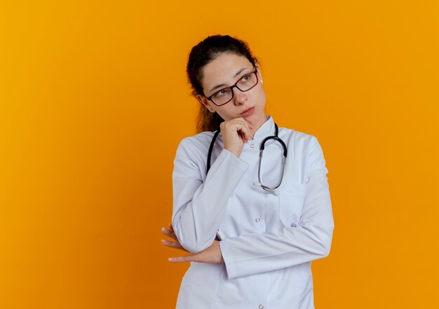 Guardando il lato pensando giovane dottoressa che indossa abito medico e stetoscopio con gli occhiali mettendo la mano sul mento isolato