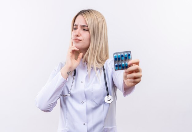 Guardando il lato pensiero medico giovane ragazza bionda che indossa stetoscopio e abito medico tenendo le pillole mise la mano sul mento sul muro bianco isolato