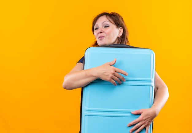 Guardando il lato soddisfatto donna di mezza età viaggiatore abbracciò la valigia sulla parete gialla isolata