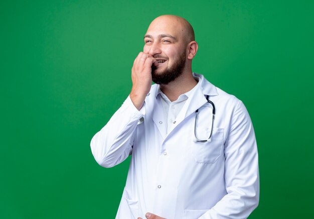 Guardando il lato soddisfatto giovane medico maschio che indossa abito medico e stetoscopio morde le unghie isolate su sfondo verde