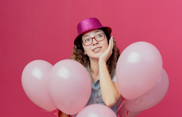 Guardando il lato soddisfatto ragazza giovane con gli occhiali e cappello rosa in piedi dietro i palloncini e mettendo la mano sulla guancia isolato su sfondo rosa