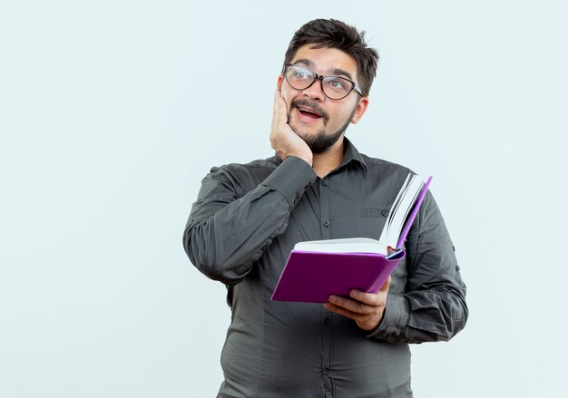 Guardando il lato sorridente giovane imprenditore con gli occhiali in possesso di libro e mettendo la mano sulla guancia isolati su sfondo bianco