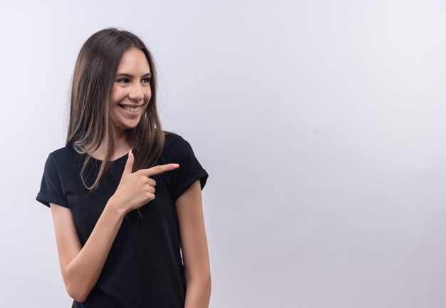 Guardando il lato sorridente giovane ragazza caucasica che indossa la maglietta nera punta a lato con il dito sul muro bianco isolato