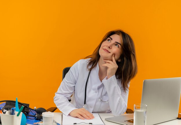 Guardando il medico femminile di mezza età soddisfatto che indossa una veste medica con lo stetoscopio seduto alla scrivania lavora sul computer portatile con strumenti medici mettendo la mano sulla guancia sulla parete arancione