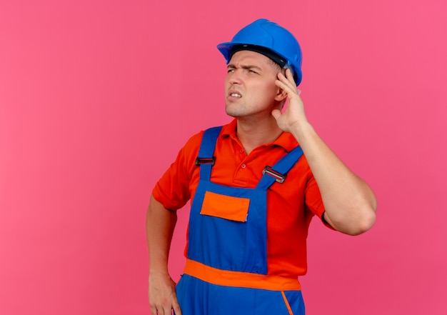 Guardando il pensiero laterale giovane costruttore maschio che indossa un casco di sicurezza e uniforme che mette la mano sull'orecchio