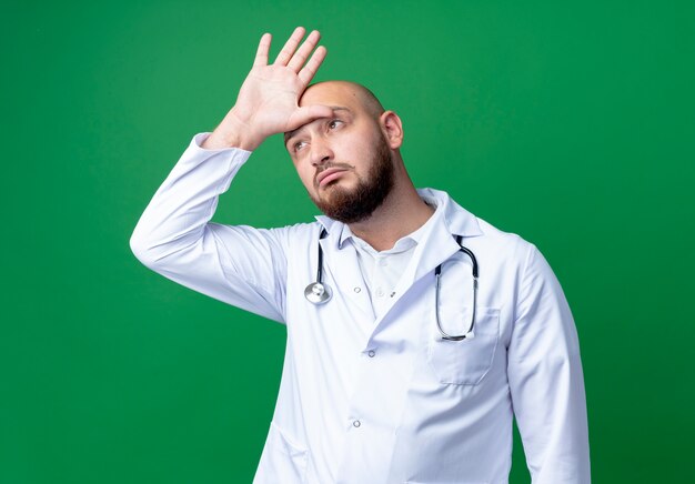 Guardando il pensiero laterale giovane medico maschio che indossa abito medico e stetoscopio mettendo la mano sulla testa isolata su sfondo verde