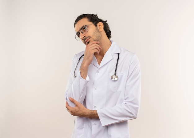 Guardando il pensiero laterale giovane medico maschio con occhiali ottici che indossa una veste bianca con lo stetoscopio che mette la mano sul mento