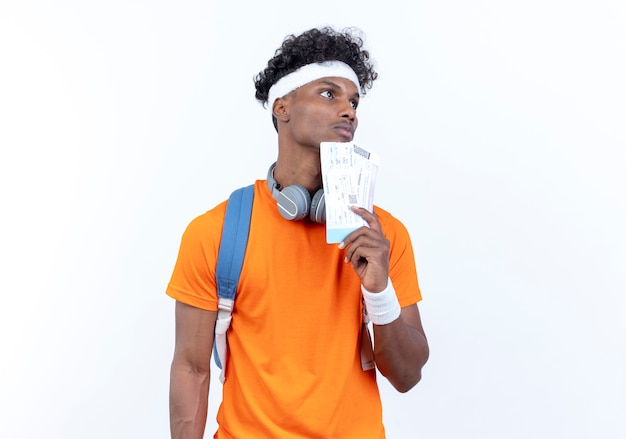 Guardando il pensiero laterale giovane uomo sportivo afro-americano che indossa la fascia e il braccialetto che tengono i biglietti isolati su priorità bassa bianca