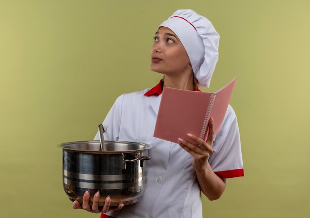 Guardando il taccuino e la casseruola della tenuta dell'uniforme del cuoco unico d'uso della femmina del cuoco del lato giovane sulla parete verde isolata