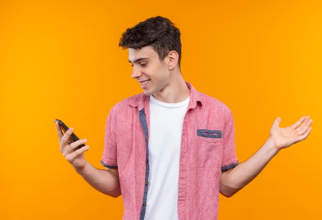 Guardando il telefono sorridente caucasico giovane uomo che indossa la camicia rosa ha alzato la mano sulla parete arancione isolata