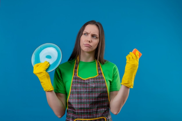Guardando l'uniforme d'uso della giovane donna di pulizia laterale in guanti che tengono plat e spugna sulla parete blu isolata