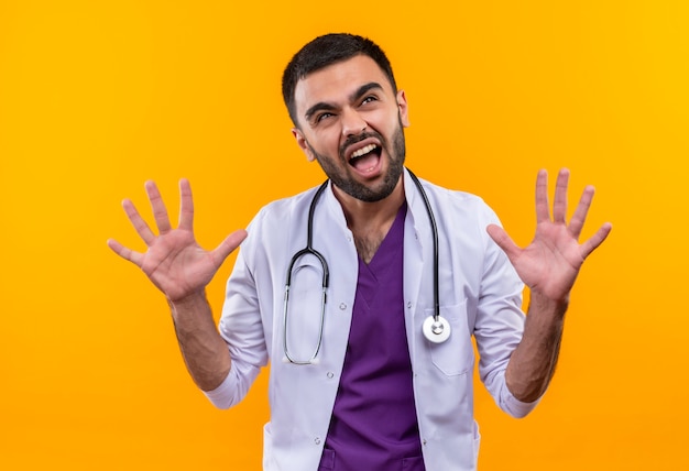 Guardando la bocca aperta del giovane medico maschio che indossa l'abito medico dello stetoscopio e alzando le mani su sfondo giallo isolato
