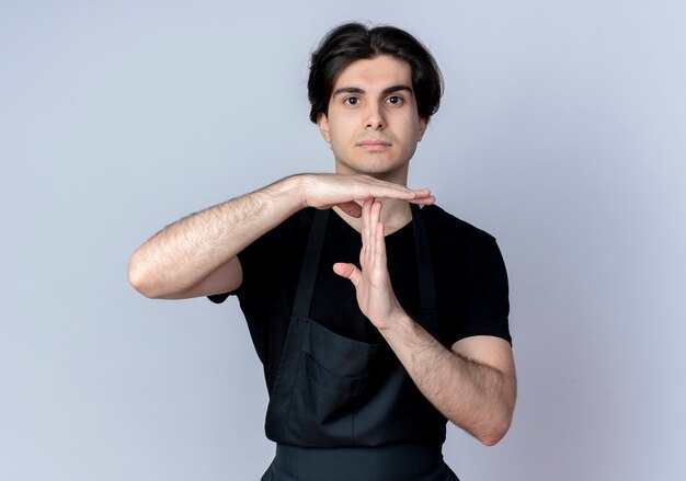 Guardando la fotocamera giovane barbiere maschio bello in uniforme che mostra il gesto di timeout isolato su priorità bassa bianca