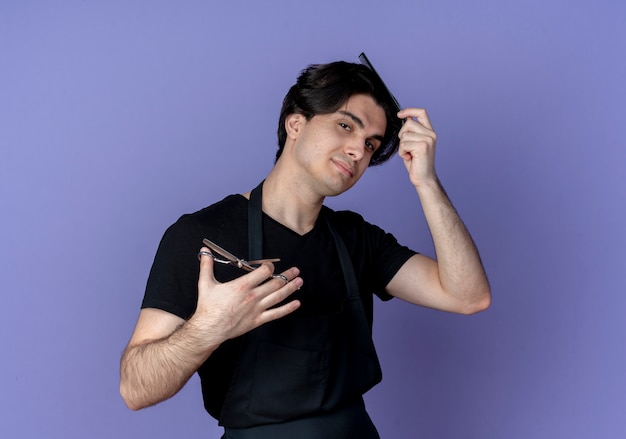 guardando la fotocamera giovane bel maschio barbiere in uniforme che pettina i capelli con il pettine e tenendo le forbici