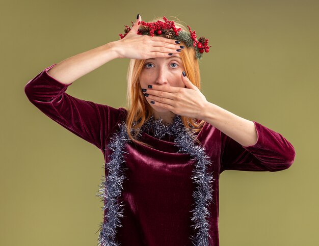 Guardando la fotocamera giovane bella ragazza che indossa un abito rosso con la corona e la ghirlanda sul collo coperto con le mani sulla fronte e la bocca isolata su sfondo verde oliva