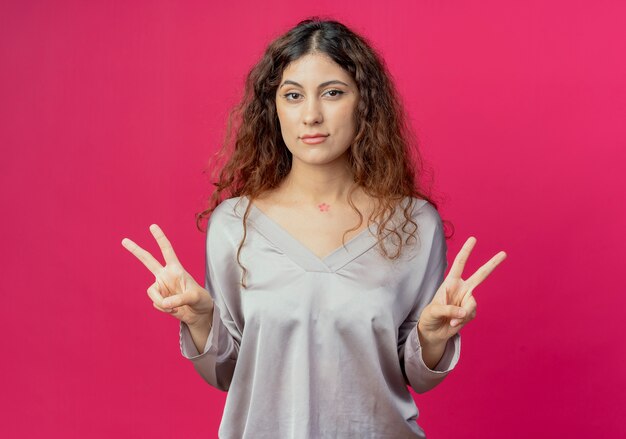 Guardando la fotocamera giovane bella ragazza che mostra il gesto di pace isolato su sfondo rosa