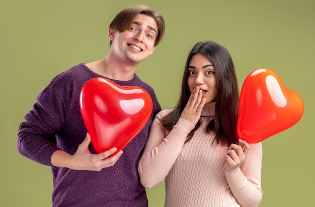 Guardando la fotocamera giovane coppia il giorno di San Valentino che tiene palloncini cuore isolati su sfondo verde oliva