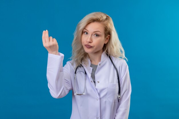 Guardando la fotocamera giovane medico che indossa uno stetoscopio in abito medico che mostra il gesto di contanti sulla parete blu