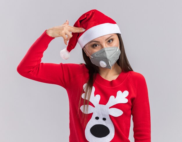Guardando la fotocamera giovane ragazza asiatica che indossa il cappello di Natale con maglione e mascherina medica che mostra il gesto della pistola isolato su sfondo bianco