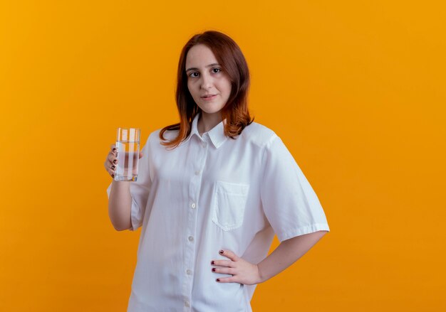 Guardando la fotocamera giovane ragazza rossa con un bicchiere d'acqua e mettendo la mano sul fianco isolato su sfondo giallo