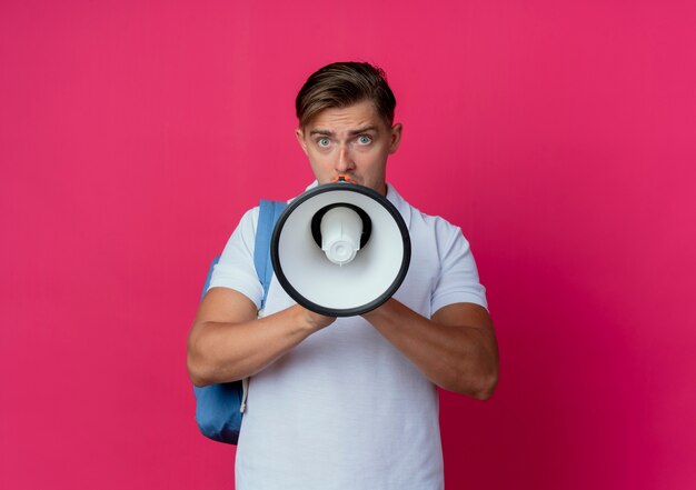 Guardando la fotocamera giovane studente maschio bello che indossa la borsa posteriore parla su altoparlante isolato su sfondo rosa