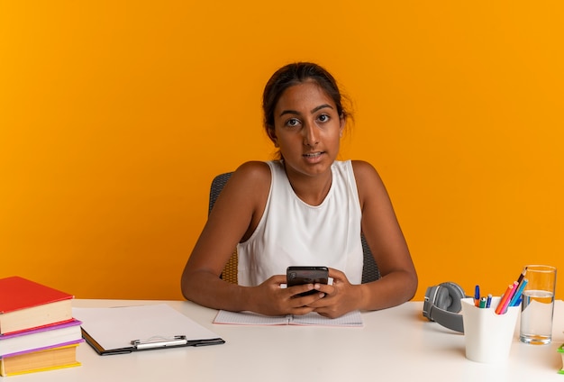 guardando la fotocamera giovane studentessa seduto alla scrivania con gli strumenti della scuola tenendo il telefono