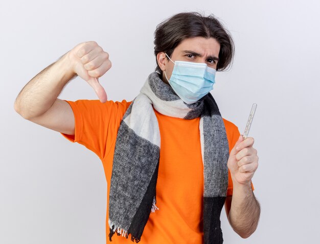 Guardando la fotocamera giovane uomo malato che indossa sciarpa e maschera medica tenendo il termometro che mostra il pollice verso il basso isolato su sfondo bianco