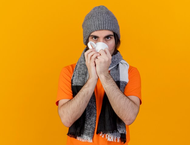 Guardando la fotocamera giovane uomo malato che indossa un cappello invernale con sciarpa bere il tè isolato su sfondo arancione