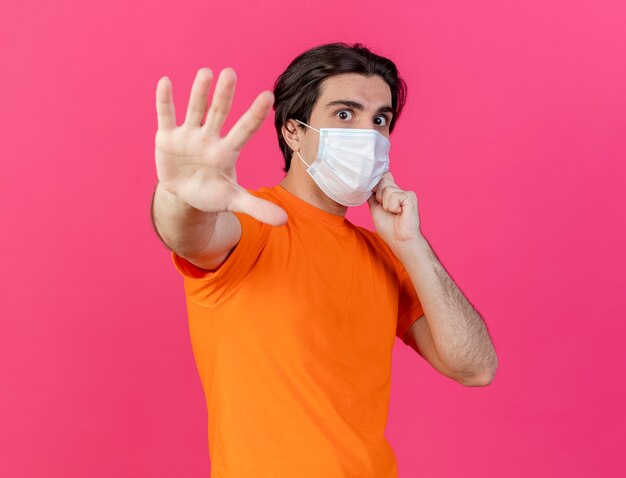 Guardando la fotocamera giovane uomo malato che indossa un cappello invernale con sciarpa e mascherina medica che mostra il gesto di arresto isolato su sfondo rosa