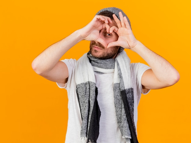 Guardando la fotocamera giovane uomo malato che indossa un cappello invernale e sciarpa che mostra il gesto del cuore isolato su sfondo giallo