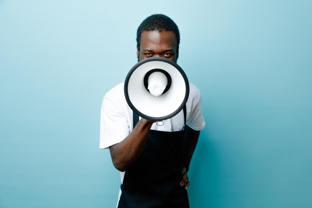 Guardando la fotocamera, il giovane barbiere afroamericano in uniforme parla con un altoparlante isolato su sfondo blu