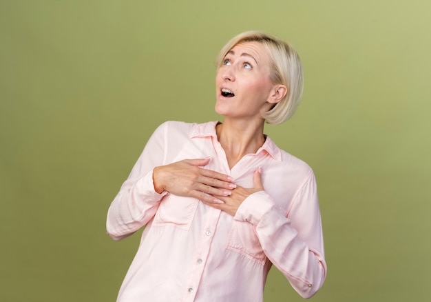 Guardando la giovane donna slava bionda spaventata che mette le mani sul cuore isolato sulla parete verde oliva