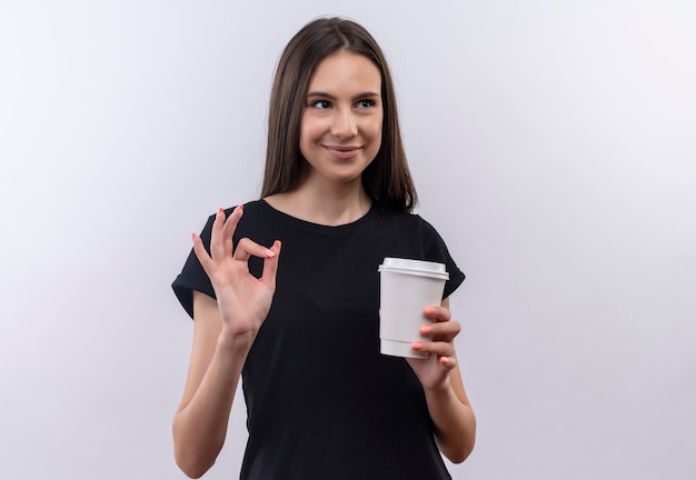 Guardando la giovane ragazza caucasica laterale che porta la tazza di caffè della tenuta della maglietta nera che mostra il gesto giusto su fondo bianco isolato