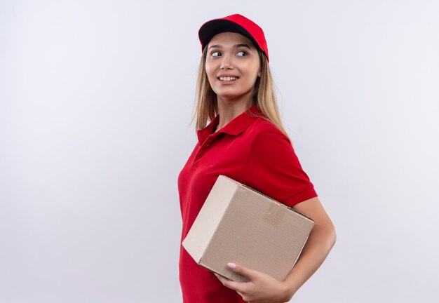 Guardando la giovane ragazza sorridente di consegna laterale che porta l'uniforme rossa e la scatola della tenuta del cappuccio isolata su fondo bianco con lo spazio della copia