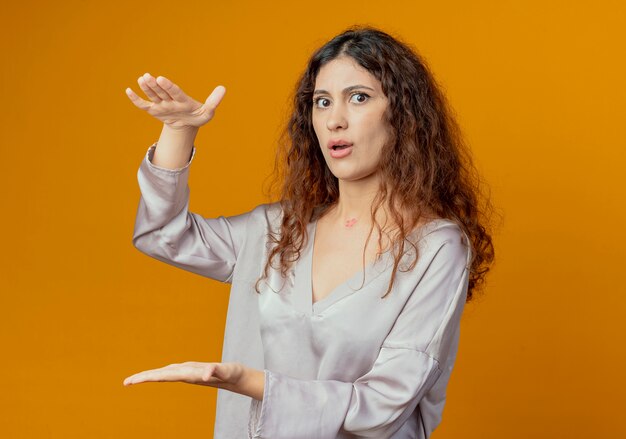 Guardando la telecamera confusa giovane ragazza graziosa che mostra le dimensioni isolate su sfondo giallo