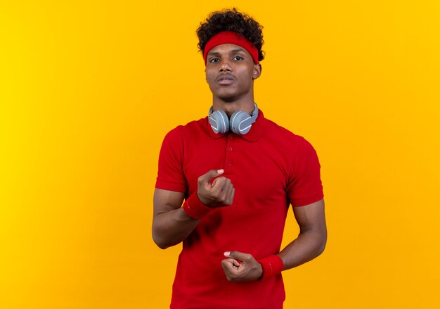 Guardando la telecamera giovane afro-americano sportivo uomo che indossa la fascia con le cuffie sul collo e polsino stringendo i pugni isolati su sfondo giallo