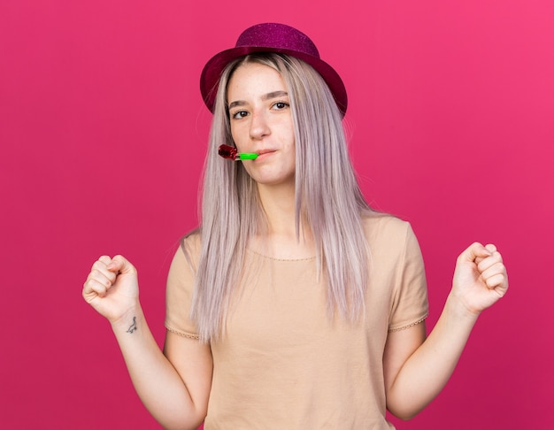 Guardando la telecamera giovane bella ragazza che indossa il cappello da festa che soffia il fischio della festa mostrando sì gesto