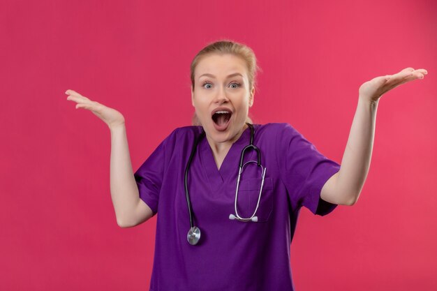 Guardando la telecamera ha sorpreso il giovane medico che indossa l'abito medico viola e lo stetoscopio allarga le mani sul muro rosa isolato