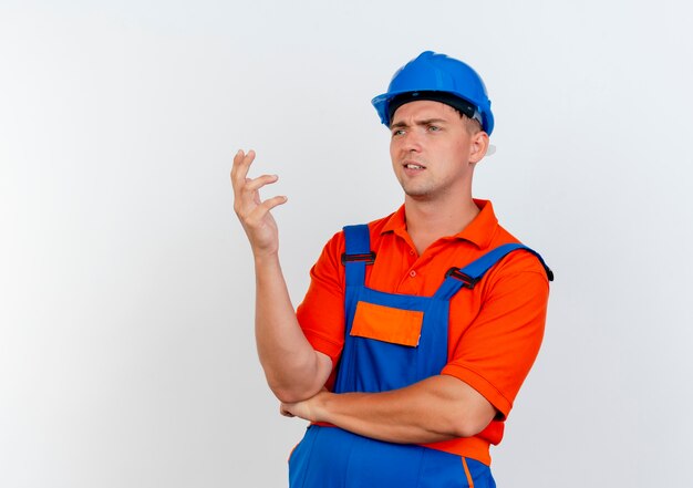 Guardando lato confuso giovane costruttore maschio che indossa l'uniforme e casco di sicurezza alzando la mano