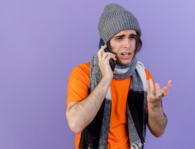 Guardando lato confuso giovane uomo malato che indossa un cappello invernale con sciarpa parla al telefono isolato su sfondo viola